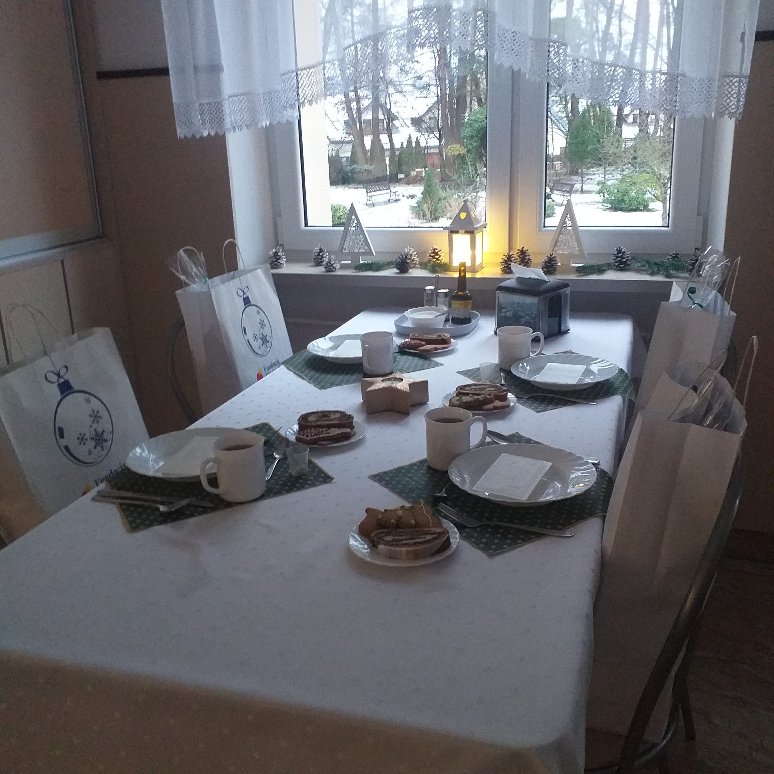 Stół nakryty białym obrusem z zastawą stołową. Na krzesłach leżą torebki prezentowe. Za oknem widać ogród. 