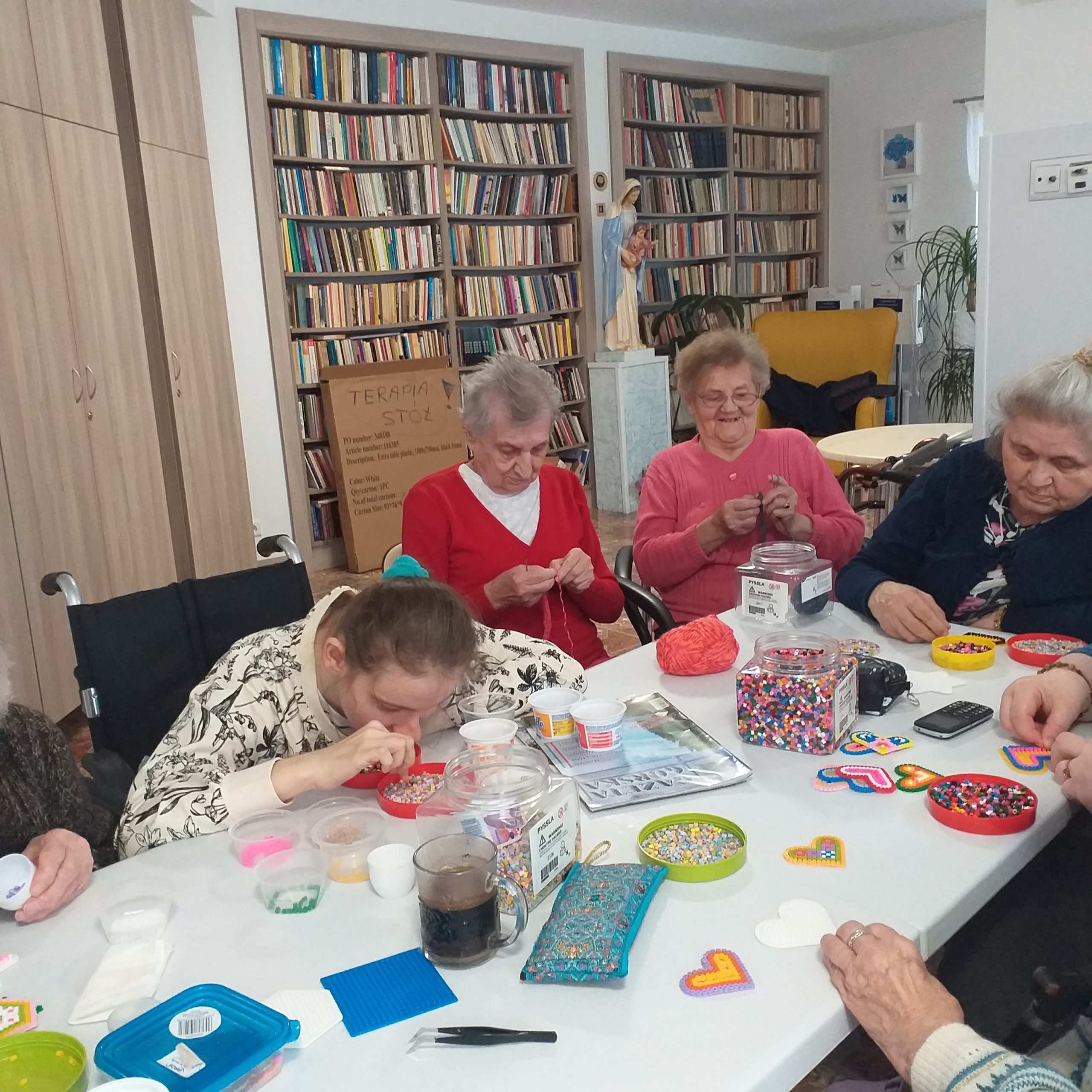 Zajęcia plastyczne. Przy stole siedzą kobiety, które wykonują ozdoby. W tle biblioteka