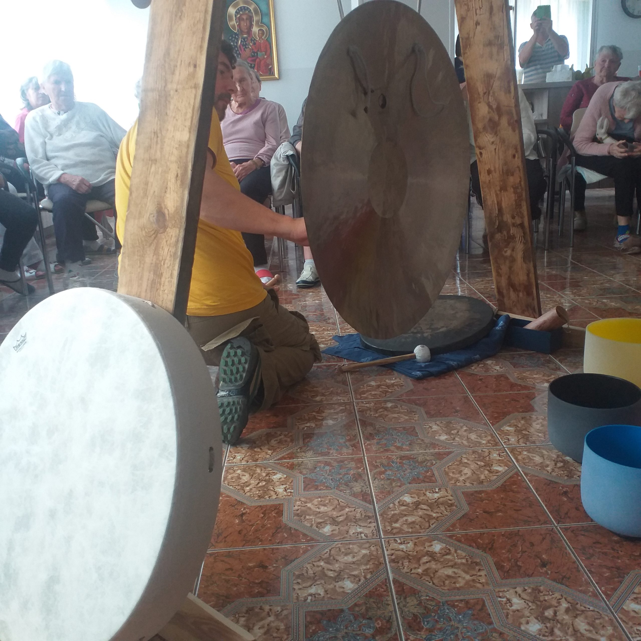 Drewniana konstrukcja z zamocowanym gongiem. Na drugim planie mieszkańcy DPS