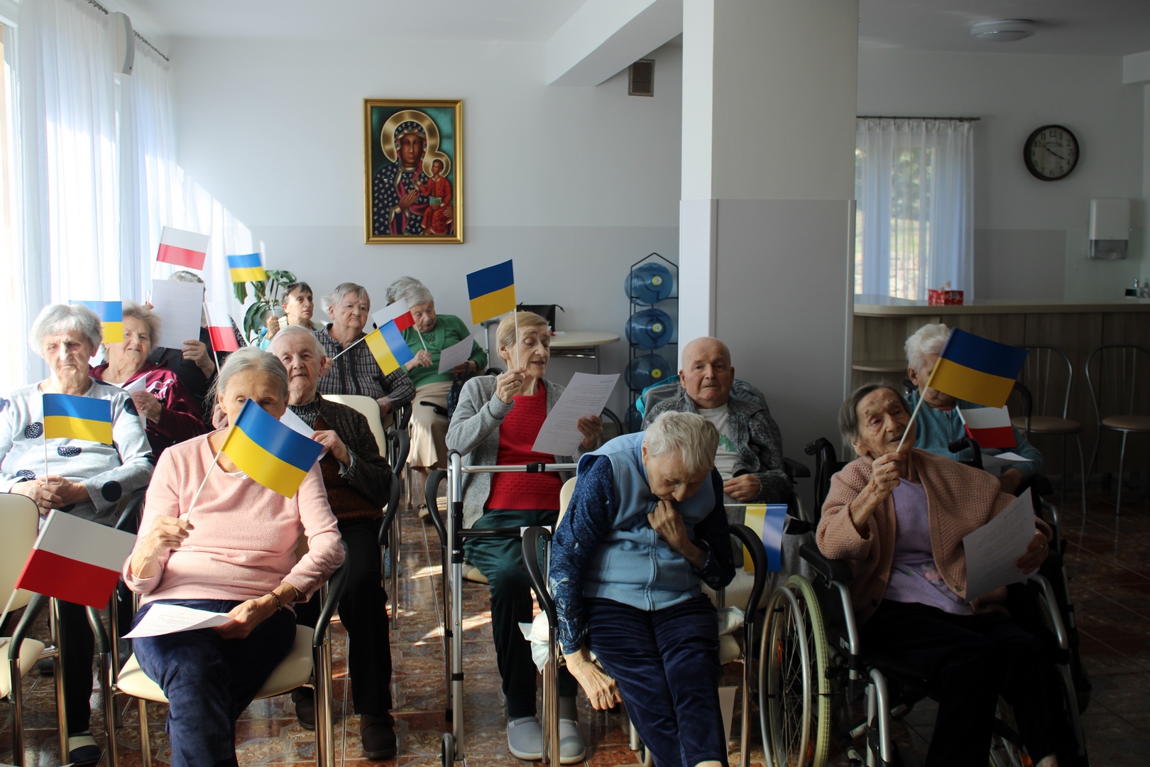 Na zdjęciu grupa 14 mieszkańców siedzących w Sali. Niektóre osoby mają na patyku flagę Ukrainy oraz Polski.