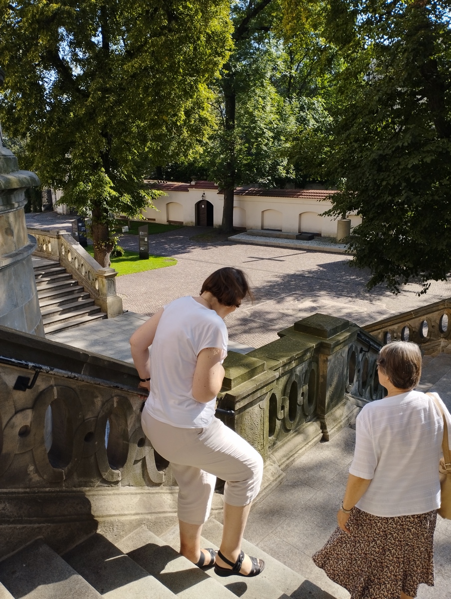 Dwie kobiety schodzące po kamiennych schodach. W oddali drzewa i mur.