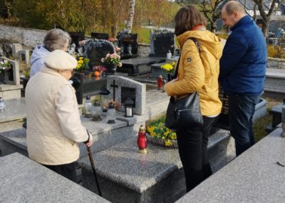 4 osoby stoją wokół nagrobku na cmentarzu. Wszyscy spoglądają na palący się znicz postawiony na grobie.