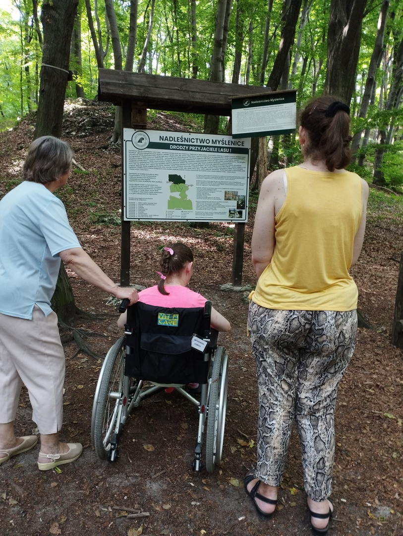 Trzy kobiety, jedna siedzi na wózku inwalidzkim w parku oglądają tablicę informacyjną nadleśnictwa Myślenice.