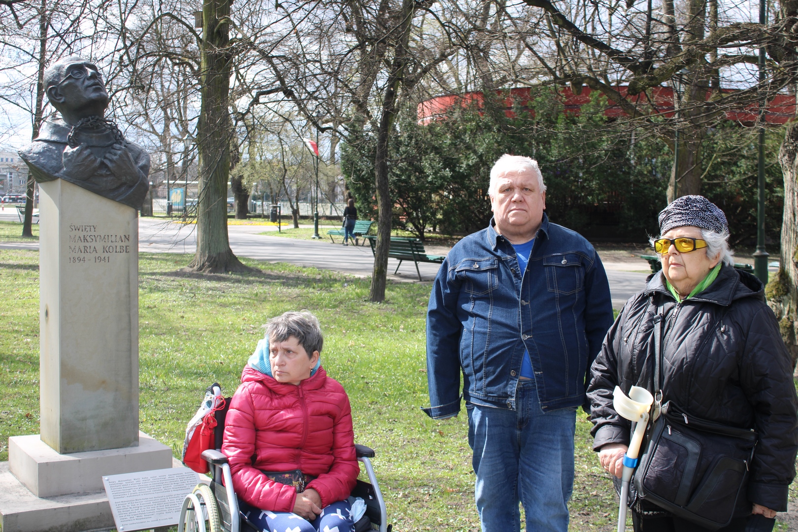 Dwie kobiety i mężczyzna przy popiersiu Św. Maksymiliana Maria Kolbe. Jedna kobieta siedzi na wózku, druga trzyma w ręku kulę.