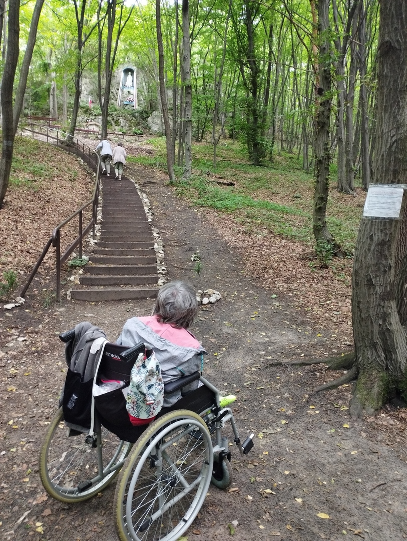 Kobieta siedząca na wózku inwalidzkim w lesie przed stromymi schodami prowadzącymi do kaplicy położonej na górze.