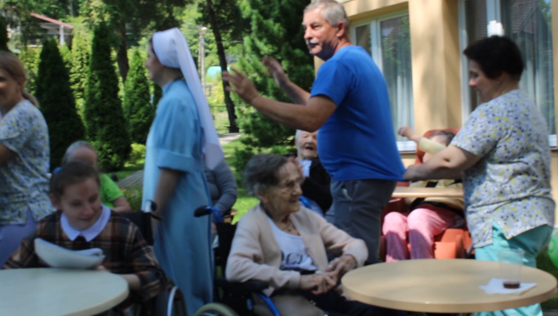 W ogrodzie przy rozłożonych stolikach siedzą kobiety, między stolikami stoją osoby, które poruszają rękami jakby tańczyły.