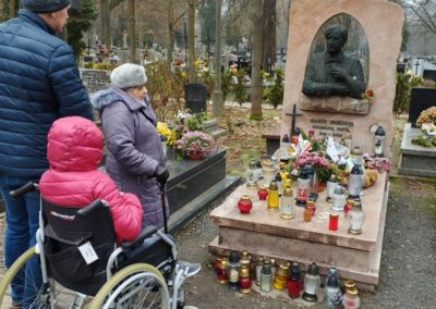 Brązowy grób Marka Grechuty. Na płycie nagrobnej złożone znicze oraz wiązanki. Przed grobowcem trzy osoby, jedna siedzi na wózku inwalidzkim.