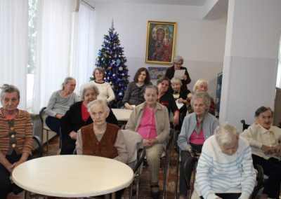 Na Sali przy okrągłych stolikach siedzą mieszkańcy DPS, wszyscy uśmiechają się do aparatu. W oddali na końcu Sali obraz i choinka.