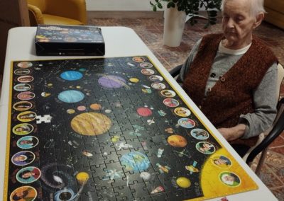 Kobieta siedzi przy dużym stole a na nim ułożone puzzle, na których widać planety, kosmos i odkrywców. Obok puzzli położona jest krzyżówka i długopis.
