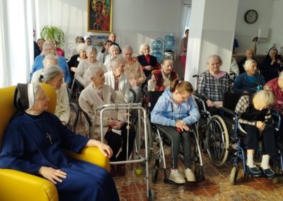 Mieszkańcy DPS siedzą w Sali po lewej stronie w żółtym fotelu siedzi siostra zakonna. Wszyscy z uśmiechem spoglądają w lewo.