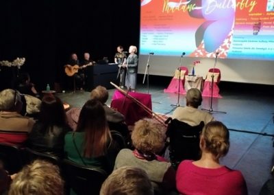 Zdjęcie sceny siedzących ludzi. Na scenie dwoje ludzi wypowiadają się do mikrofonu.