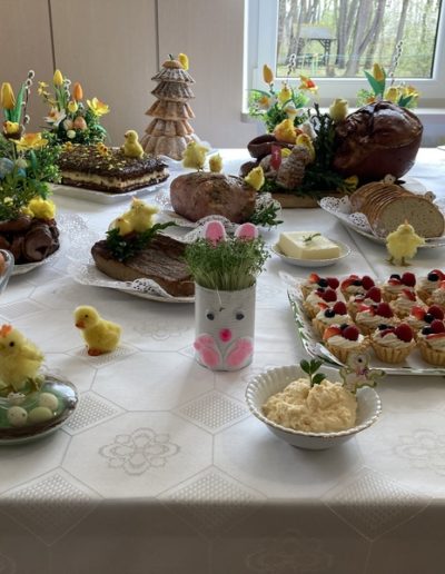 Wielkanocny stół przykryty białym obrusem a na nim poukładane pisklaki, pisanki, chleb, jaja i pisanki, babeczki, ciasta. Wszystko udekorowane żółtymi tulipanami.