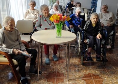 grupa mieszkańców siedzi na sali. Pośrodku nich wazon z papierowymi kwiatami.