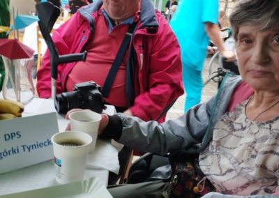 Kobieta i mężczyzna siedzą przy stoliku. Na stole kawa, kromka chleba ze smalcem, aparat fotograficzny oraz napis "DPS Podgórki Tyniecki