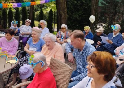 Grupa mieszkańców siedzi w ogrodzie przy stolikach. Część z nich mają założone kolorowe maski.