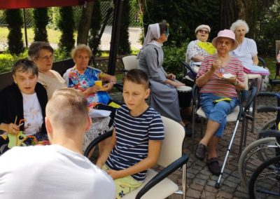 Grupa mieszkańców siedzi w ogrodzie przy stolikach i rozmawiają z gośćmi.