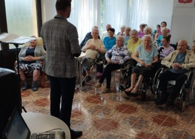 Sala konferencyjna na środku niej Mężczyzna w marynarce. Mieszkańcy siedzą przed nim i patrzą się na mężczyznę.