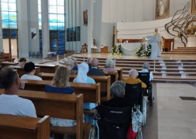 Mieszkańcy DPS uczestniczą we Mszy Świętej. Siedzą w ławkach, w oddali widać kapłana przy ołtarzu.