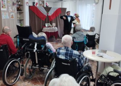 W sali mieszkańcy obserwują spektakl dotyczący niepodległości. W oddali na ściance wisi flaga RP