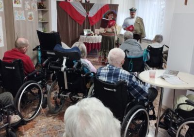 W sali mieszkańcy obserwują spektakl dotyczący niepodległości. W oddali na ściance wisi flaga RP