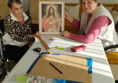 Dwie mieszkanki siedzą przy stole i w rękach trzymają obraz Pana Jezusa wyszywany.