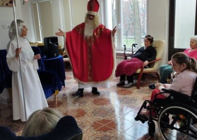 Pośrodku sali stoi z rozłożonymi rękami Święty Mikołaj i spogląda na mieszkańców. Po jego prawej stoi aniołek.