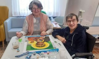 7.03.25 IMG-20250218-WA0004s Na zdjęciu znajdują się dwie osoby siedzące przy stole, pracujące nad kolorową układanką lub dziełem sztuki. Na stole rozłożone są różne przybory artystyczne i kawałki układanki. W tle widać kanapę oraz okno z zasłonami.
